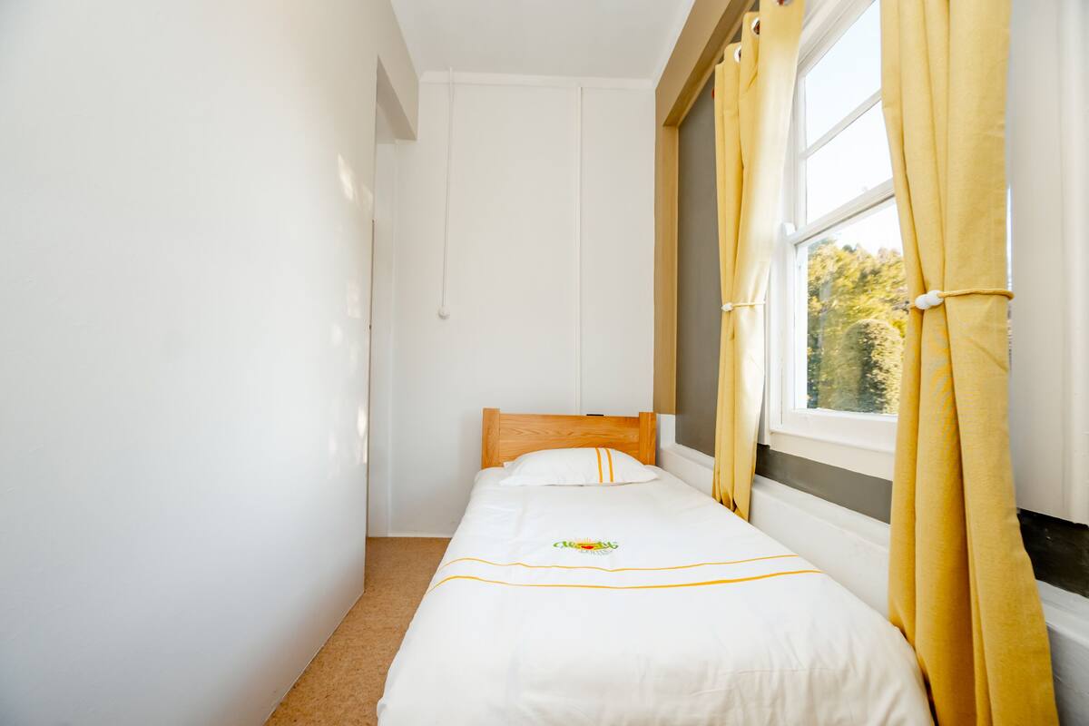 A small, single-bed room with a wooden headboard, a window with yellow curtains, and a white bedspread with a small embroidered design.