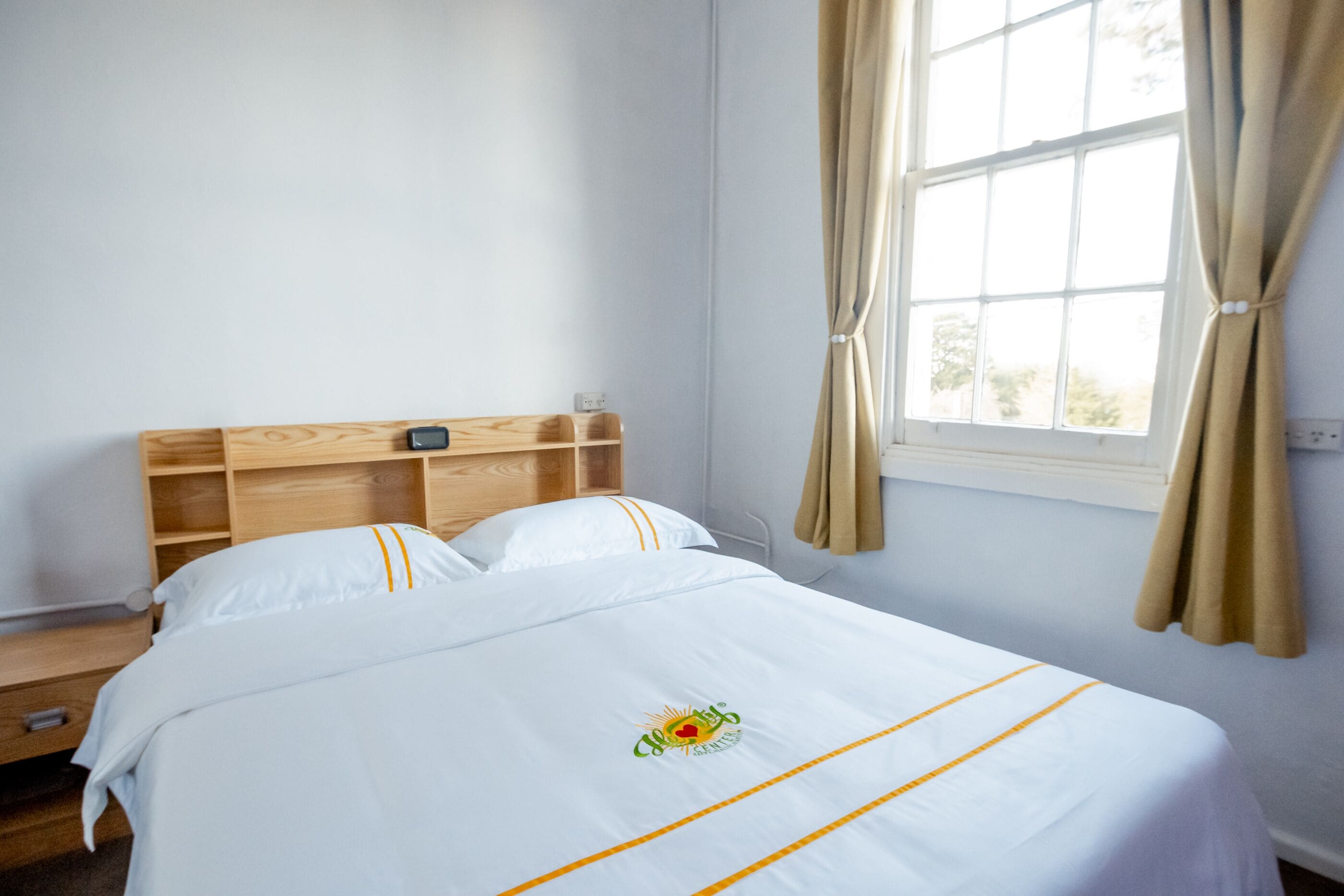 A neatly made bed with a logo, a wooden headboard, and a window with curtains.