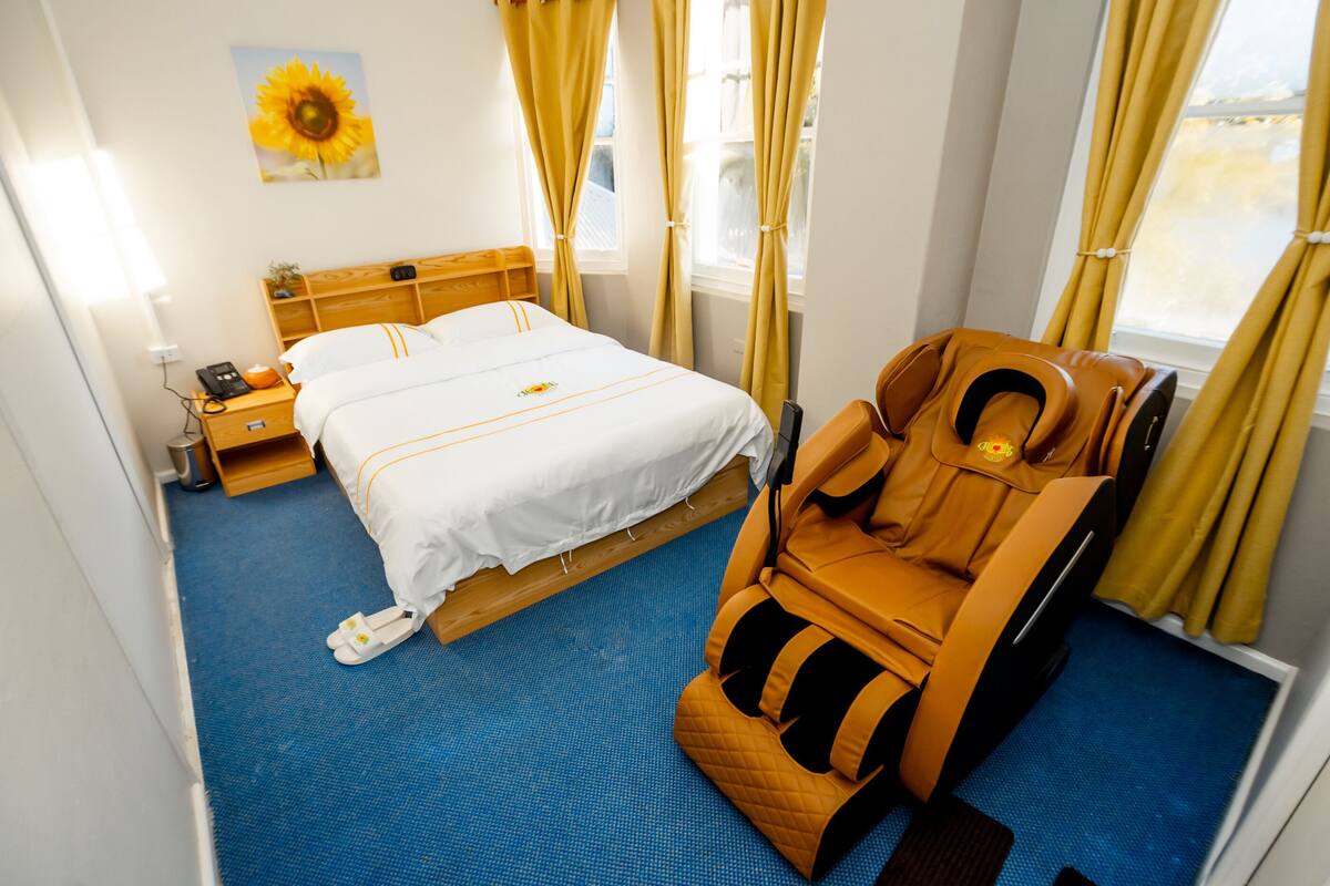 A hotel room with a bed, a massage chair, a bedside table with a phone, and a window with curtains.