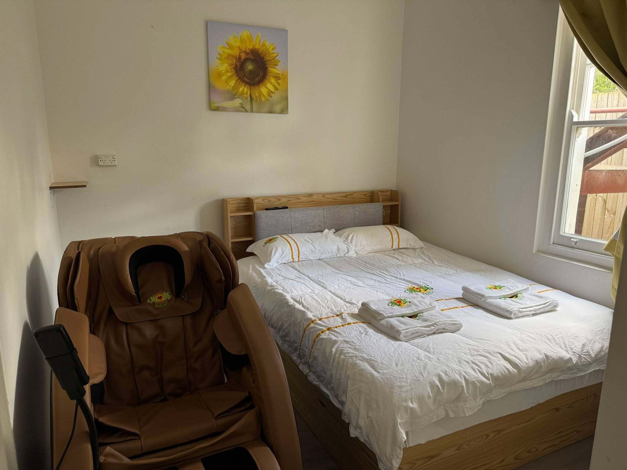 A bedroom with a bed, a massage chair, a painting of a sunflower, and a window with curtains.