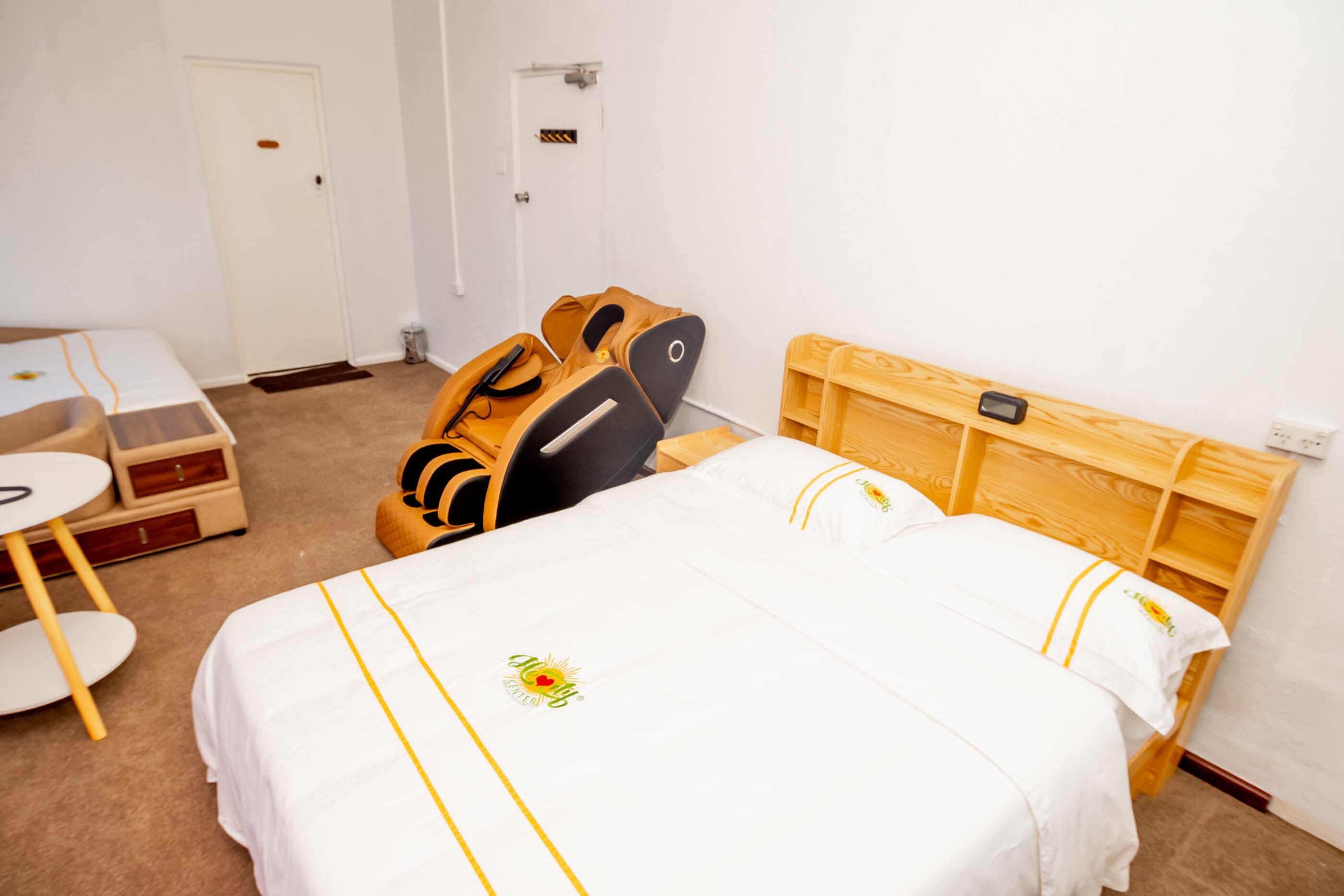 A hotel room with a bed, a massage chair, a small table, and a wooden headboard.