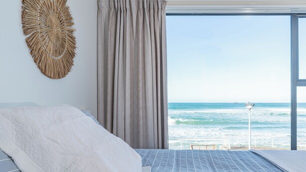 A bedroom with a large bed, a view of the ocean, and a beach visible through the window. - Esplanade Apartments (Dunedin)