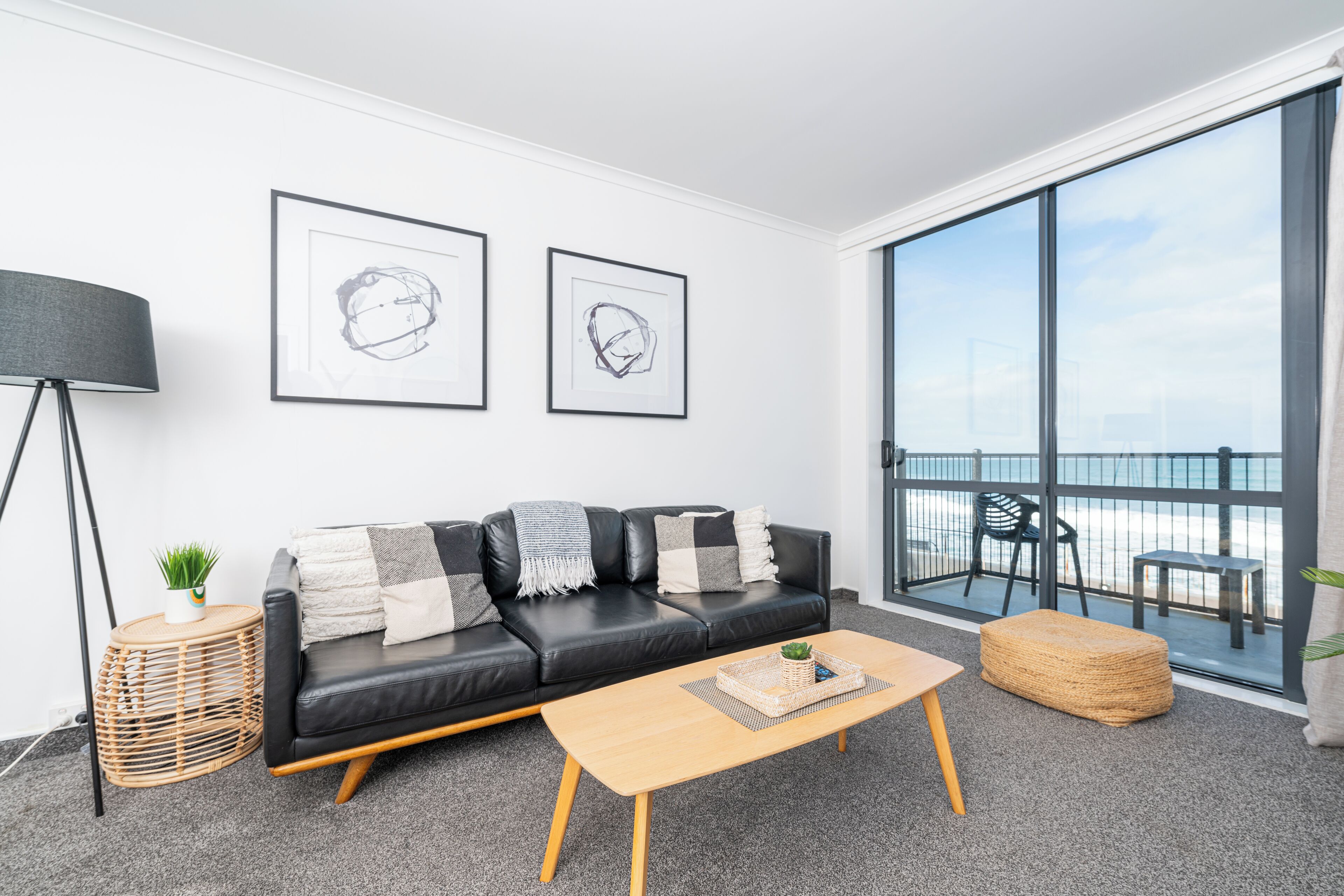 A modern living room with a black leather sofa, a wooden coffee table, and a large window offering a view of the beach.