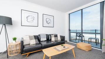 A modern living room with a black leather sofa, a wooden coffee table, and a large window offering a view of the beach.