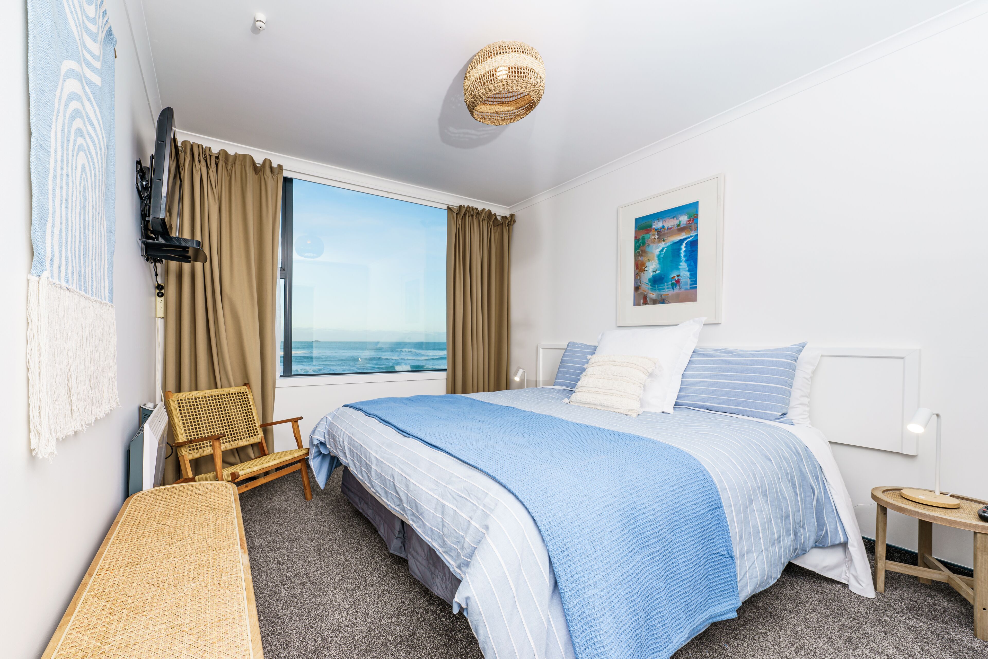 A bedroom with a large bed, a chair, a small table, and a view of the ocean.