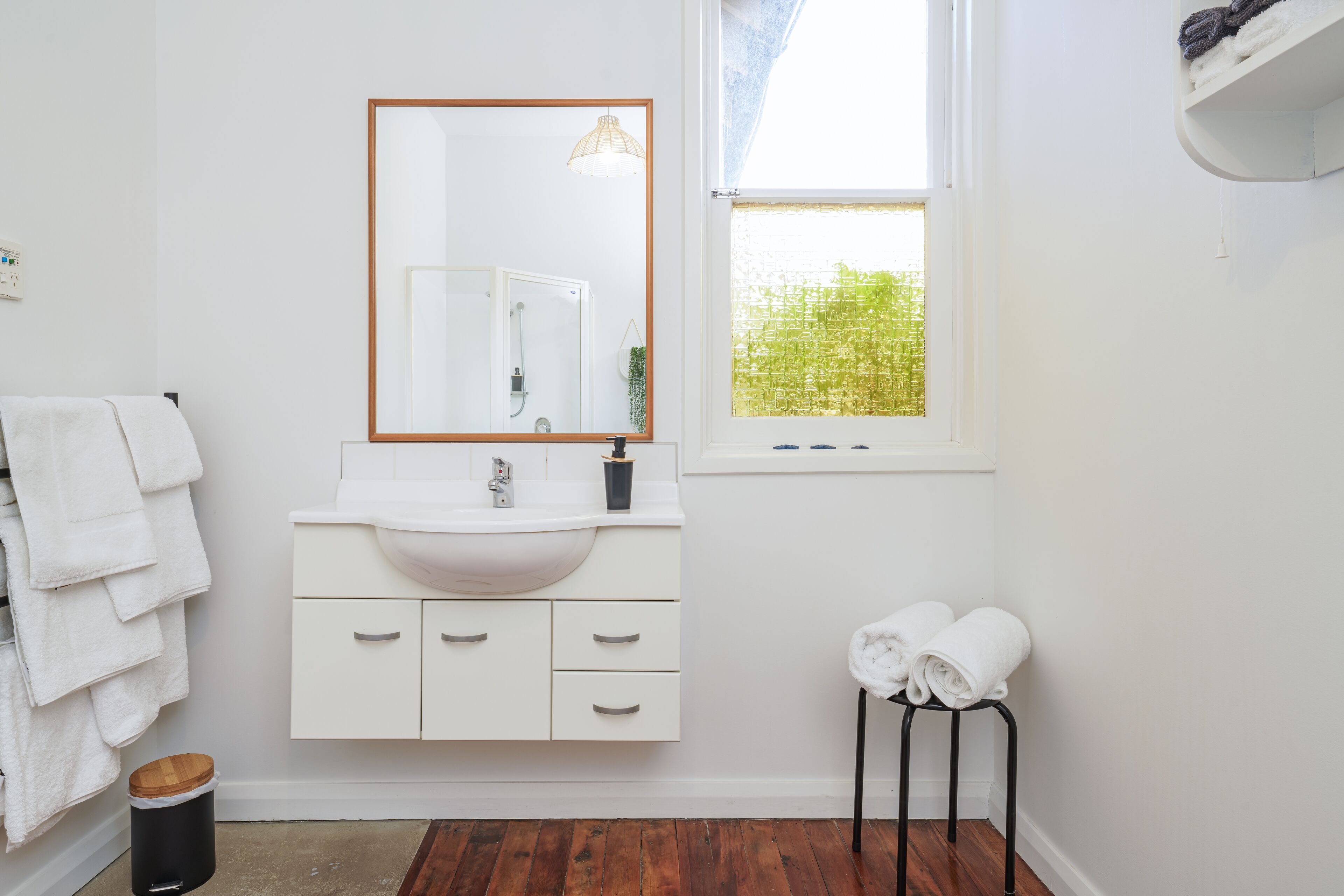 A bathroom with a white sink, mirror, and wooden floor. There are towels on a rack and a stool with towels.