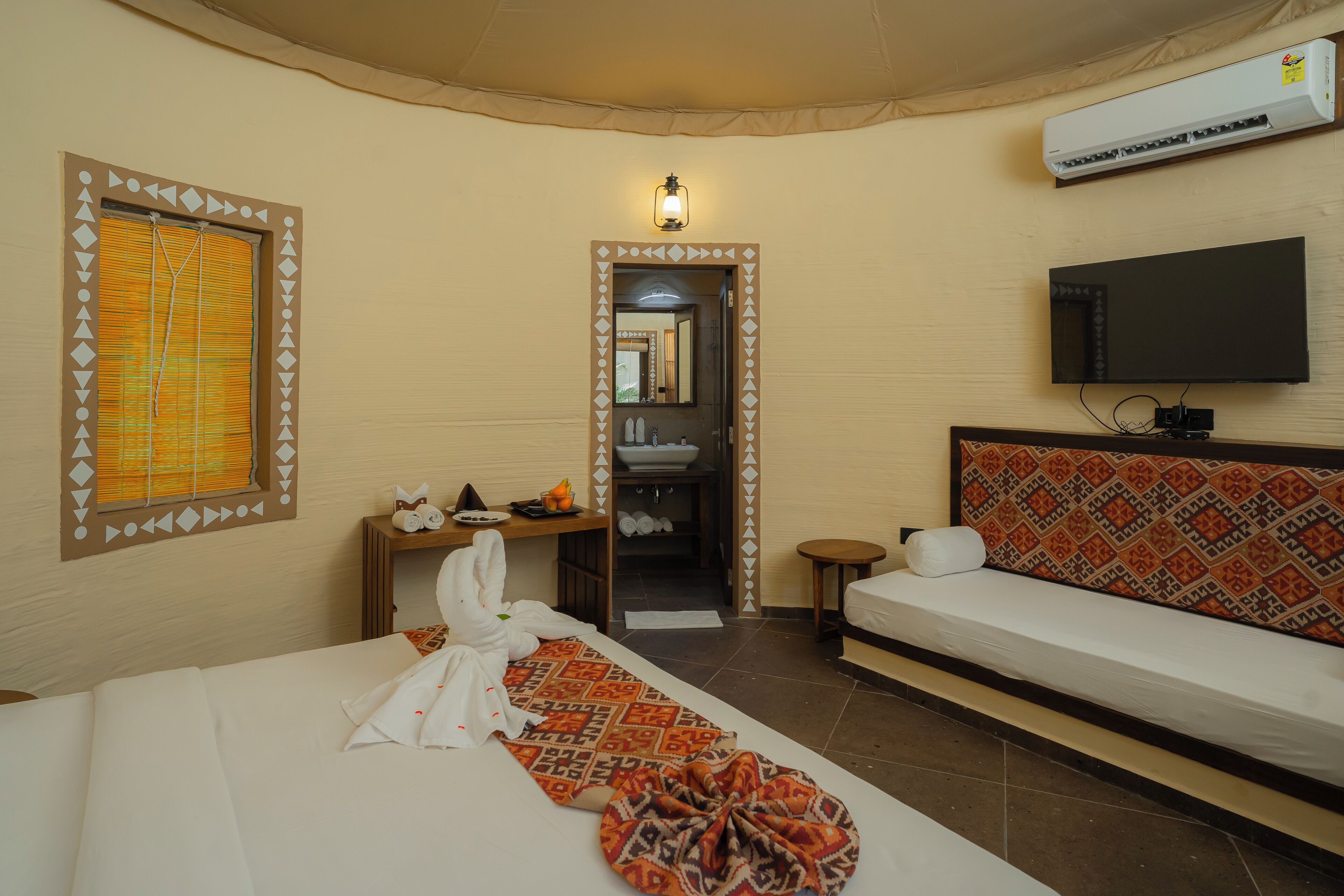 A hotel room with a bed, a sofa, a television, an air conditioning unit, and a bathroom visible through an open door.