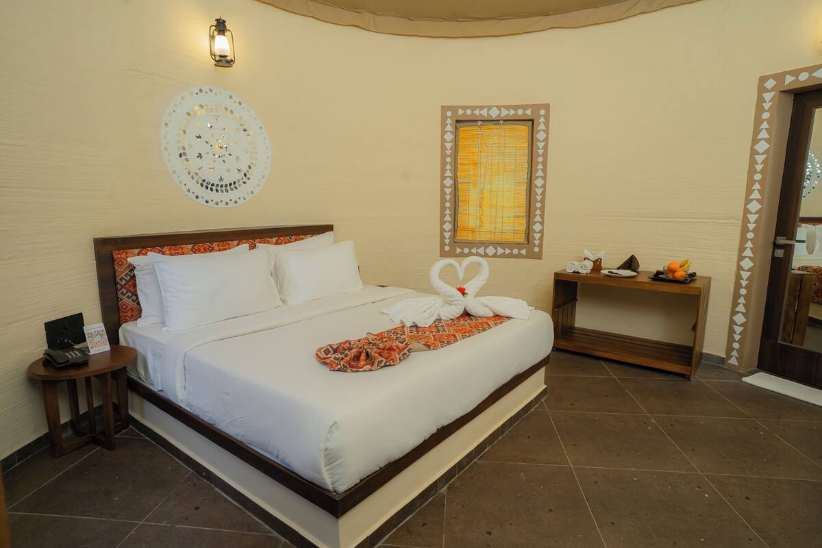 A hotel room with a large bed, a bedside table with a lamp, a wall decoration, and a window with blinds.