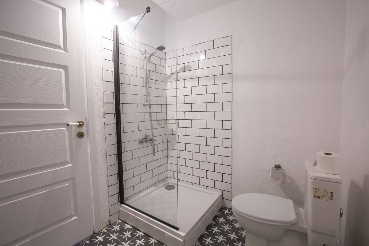A bathroom with a glass-enclosed shower, white tiled walls, a toilet, and a patterned floor.