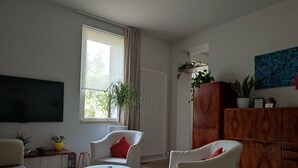Un salon avec un téléviseur, des chaises blanches, une petite table et un meuble en bois orné de plantes.