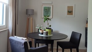 Un coin repas moderne avec une table ronde, quatre chaises et un luminaire suspendu. Deux œuvres d’art encadrées ornent le mur, et une plante en pot est posée sur la table.
