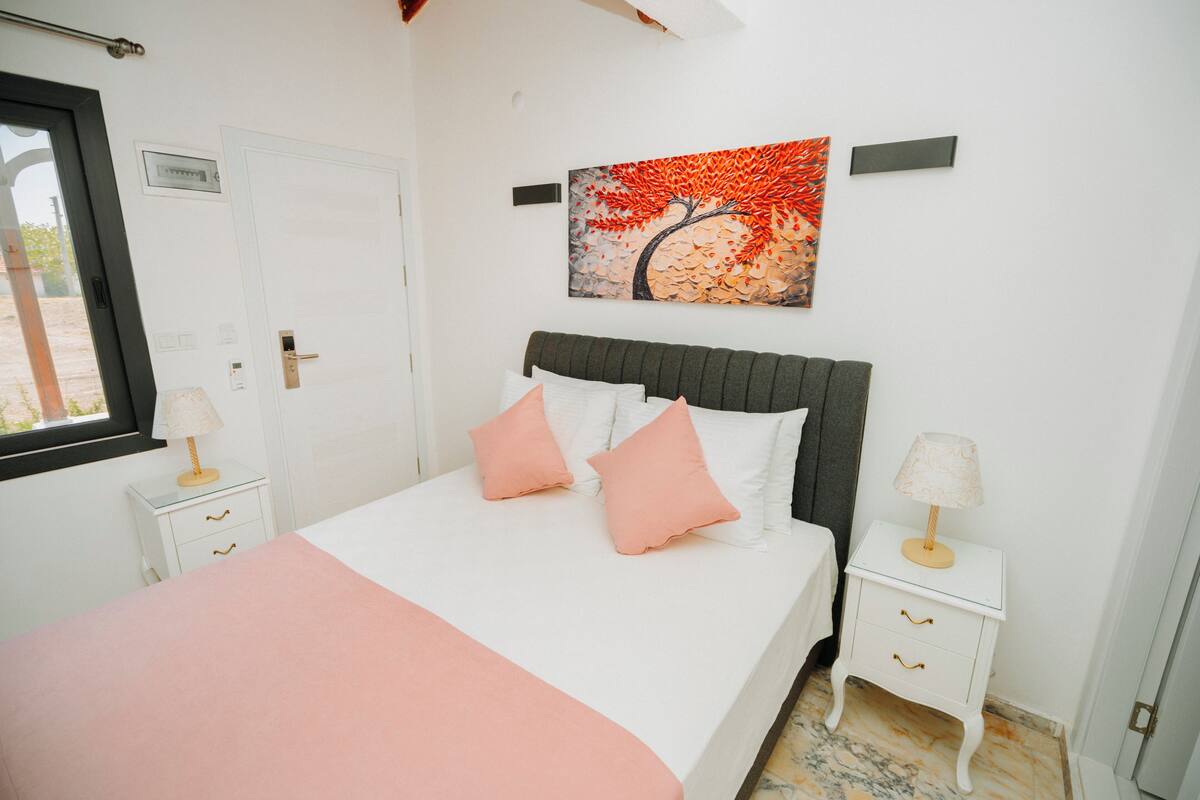 A neatly arranged bedroom with a bed, two bedside tables, a window, and a wall-mounted painting.