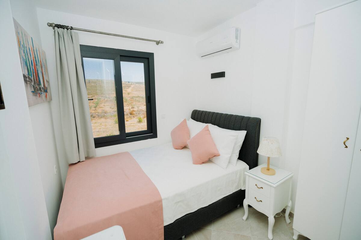 A bedroom with a bed, a nightstand, a window with curtains, an air conditioning unit, and a view of the outdoors.