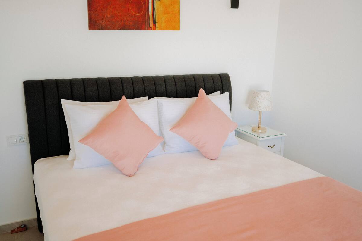 A neatly made bed with a black headboard, white and pink pillows, and a white nightstand with a lamp.