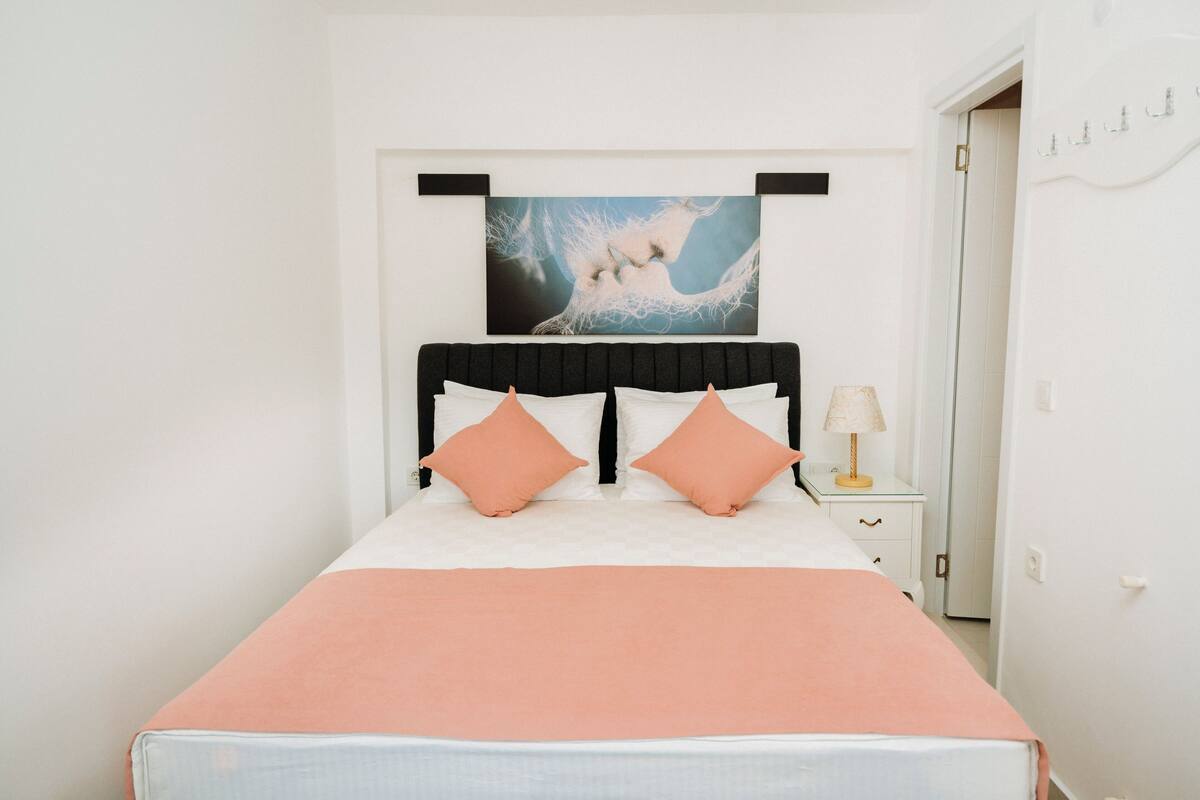 A neatly made bed with a peach-colored bedspread and two peach pillows, a black headboard, and a wall-mounted painting above.