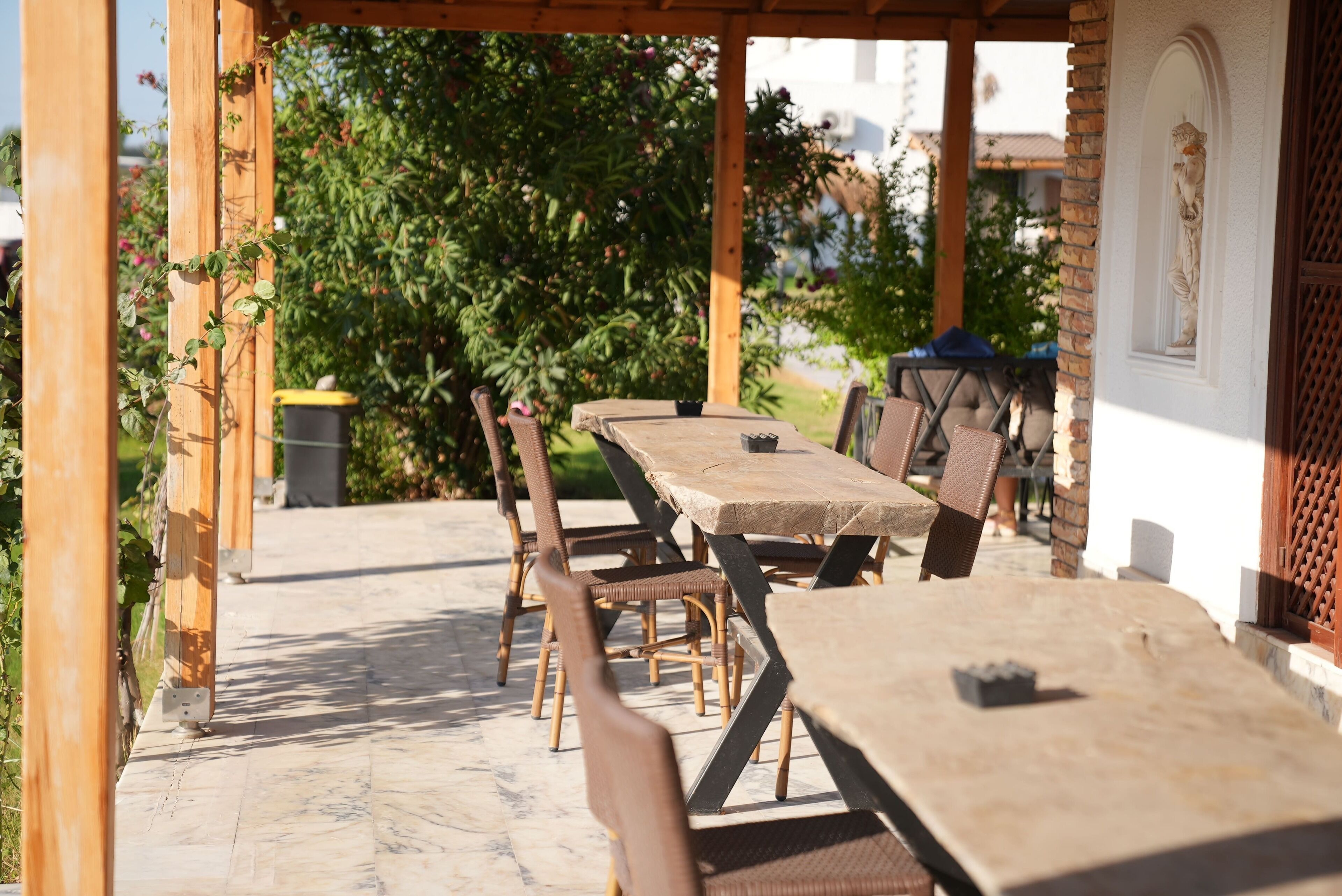Eine Außenpatio mit einer Holzpergola, einem Tisch und Stühlen, und Blick auf einen Garten sowie ein Gebäude im Hintergrund.