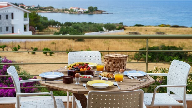 A dining table set with breakfast items, white chairs, and a view of the sea and mountains in the background. - Villa Luisa Exclusive Residences (Monemvasia)