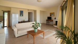 A living room with a sofa, a coffee table, a TV, a dining table, and chairs. There is a plant on the coffee table and a window with curtains. - Villa Luisa Exclusive Residences (Monemvasia)