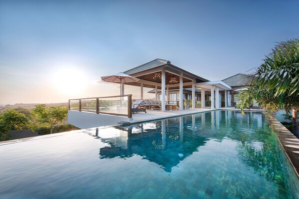 A modern infinity pool with a clear view of the horizon, a covered outdoor seating area, and a well-maintained landscape.