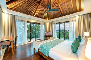 A spacious bedroom with a large bed, a desk, and a chair, featuring a ceiling fan, large windows, and a view of the outdoors.