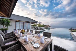 An outdoor dining area with a view of the ocean, featuring wicker furniture, neatly set tables, and a glass-bottomed infinity pool.