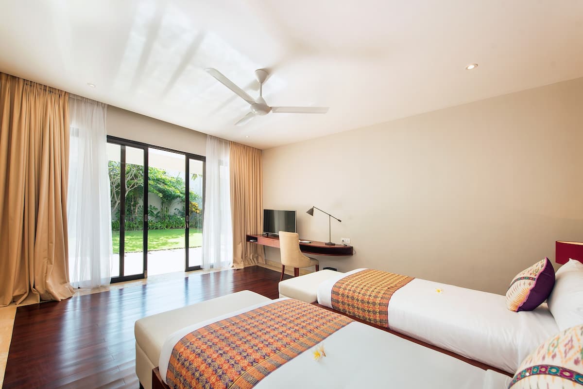 A hotel room with two beds, a desk, a chair, a ceiling fan, and large windows with curtains.