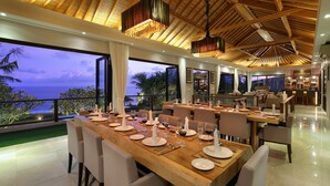 A dining area with a long table set for a meal, large windows with a view of the ocean, and a high ceiling with wooden beams. - Karang Saujana Estate (Kutuh)