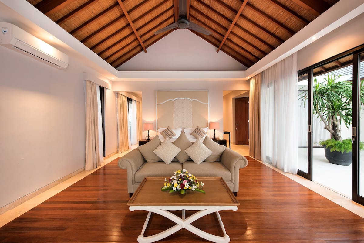 A spacious room with a wooden ceiling, a sofa with cushions, a coffee table with flowers, and a large window with a plant outside.
