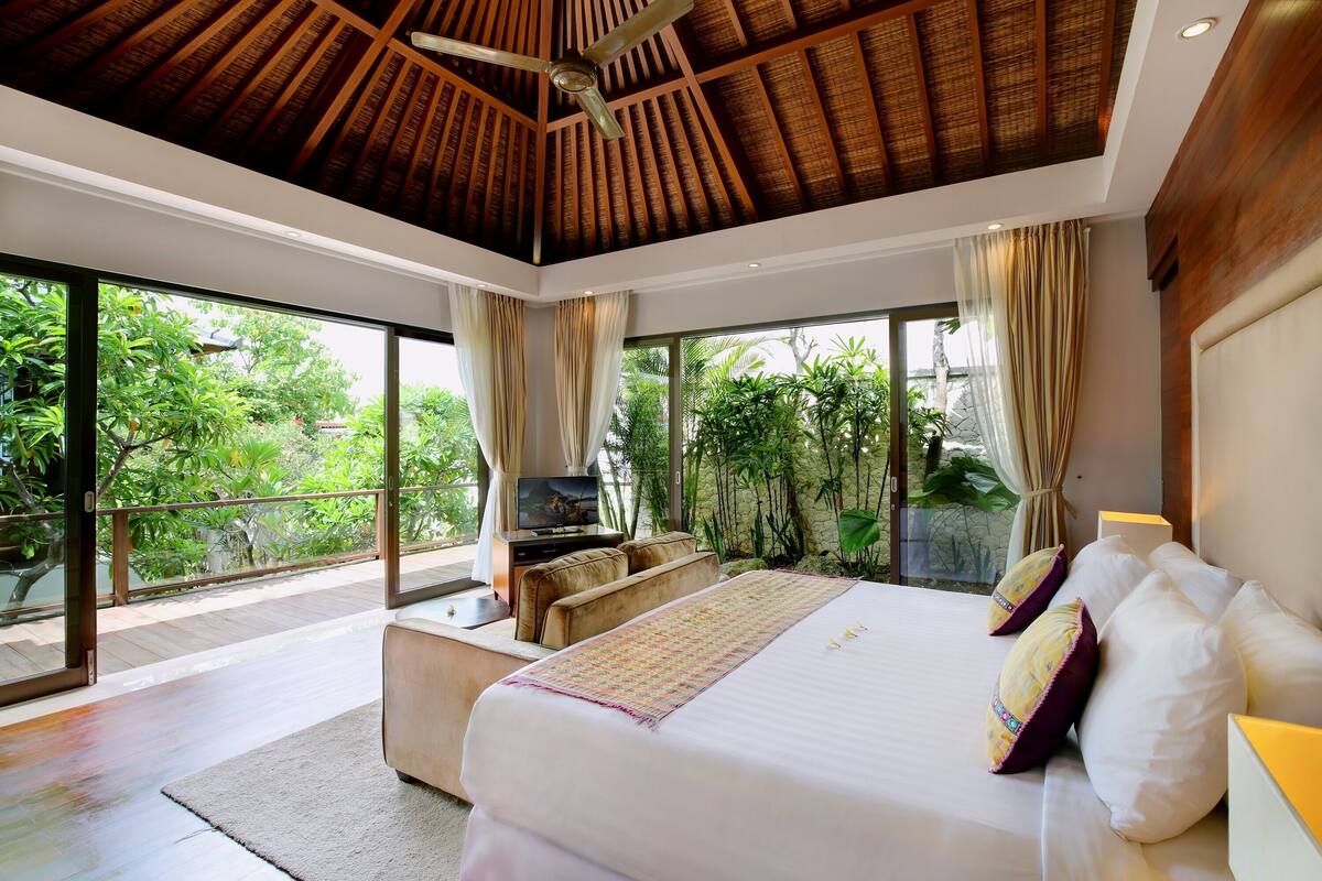 A modern bedroom with a large bed, a TV, and a balcony with a view of greenery.