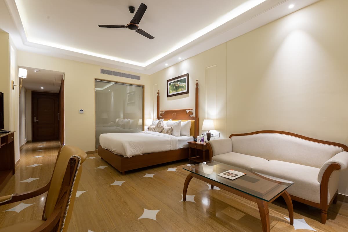 Quarto de hotel com cama, sofá, mesa de centro de vidro, cadeira, ventoinha no teto e espelho.