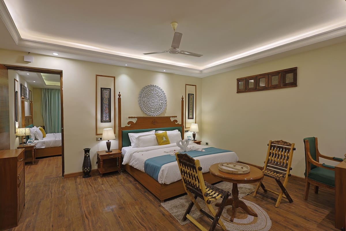 Quarto de hotel com piso de madeira, cama com colcha azul e branca, duas cadeiras de madeira, uma mesa redonda e um ventilador de teto.