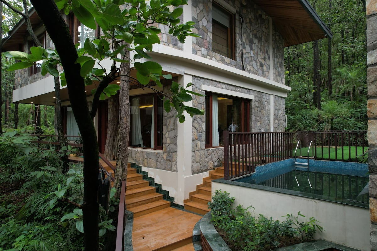 Uma casa de pedra com escadaria de madeira que dá para uma varanda, rodeada por vegetação exuberante.