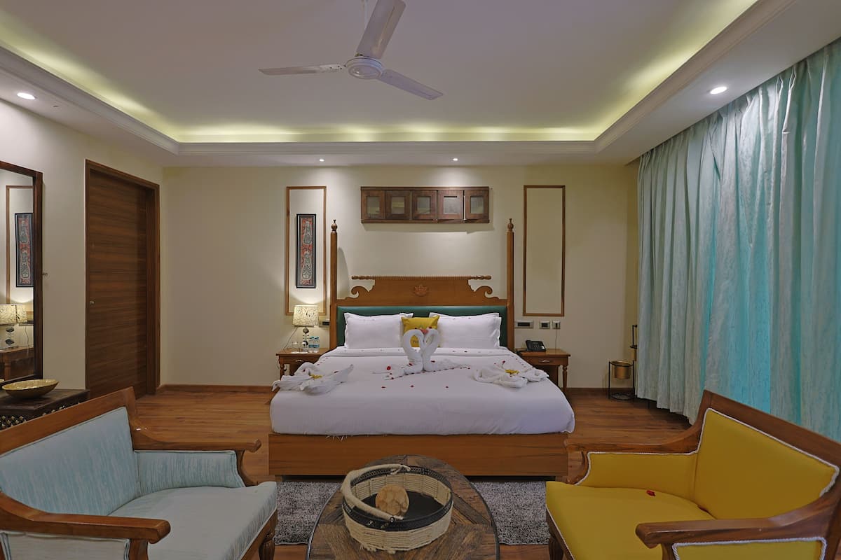 Quarto de hotel com uma cama grande, duas cadeiras de madeira, um ventilador de teto e uma mesa de cabeceira com abajur.