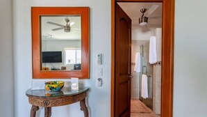 Un couloir avec une porte en bois donnant sur une salle de bain. On y trouve un miroir, au-dessus duquel est fixé un ventilateur de plafond, ainsi qu’une petite table sur laquelle se trouve un bol.