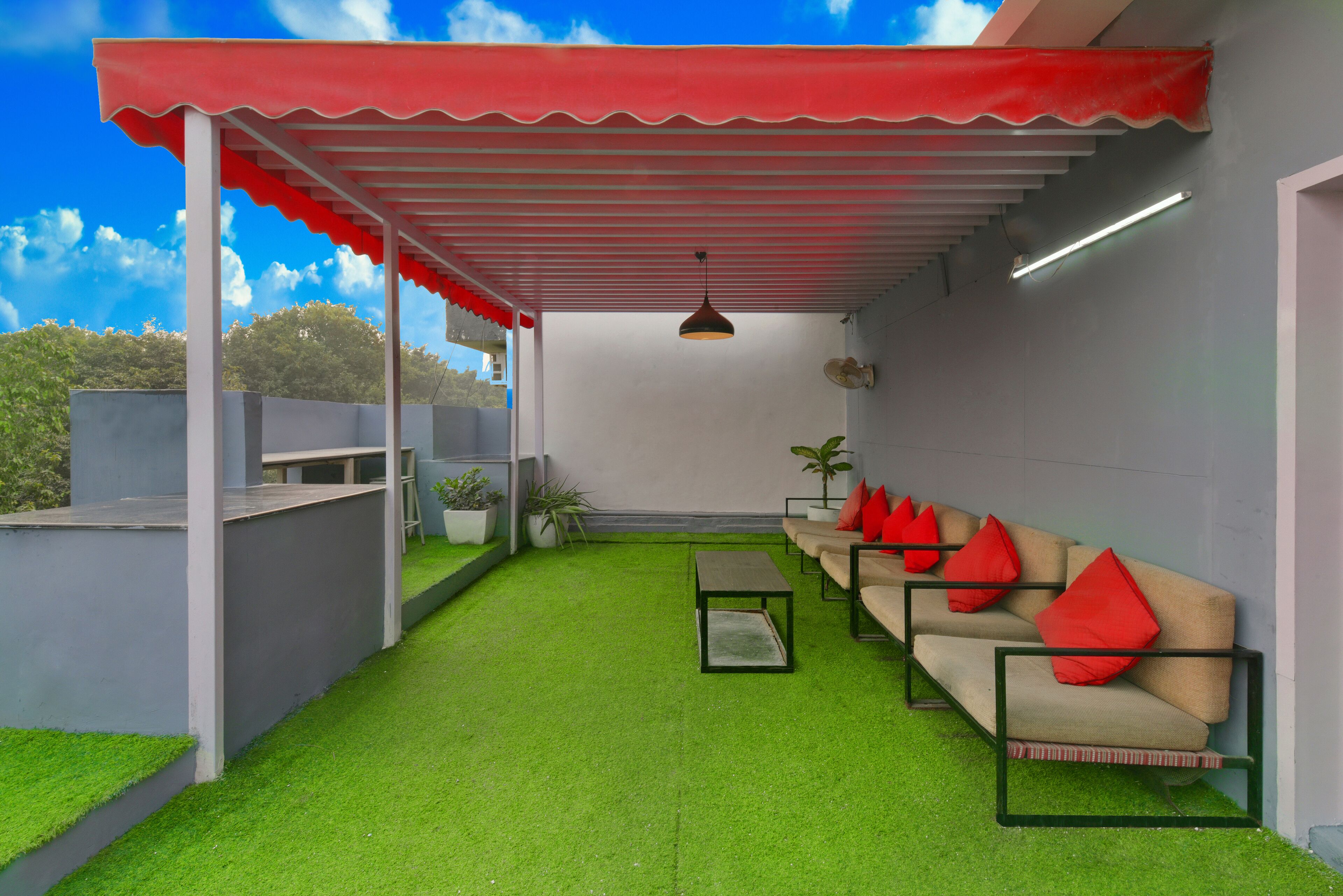 Un espace extérieur aménagé avec une zone salon couverte, recouverte d’un gazon synthétique, comprenant un canapé avec des coussins rouges et un luminaire suspendu.