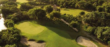 Campo de golf con búnkeres de arena, un obstáculo acuático y un horizonte urbano al fondo.