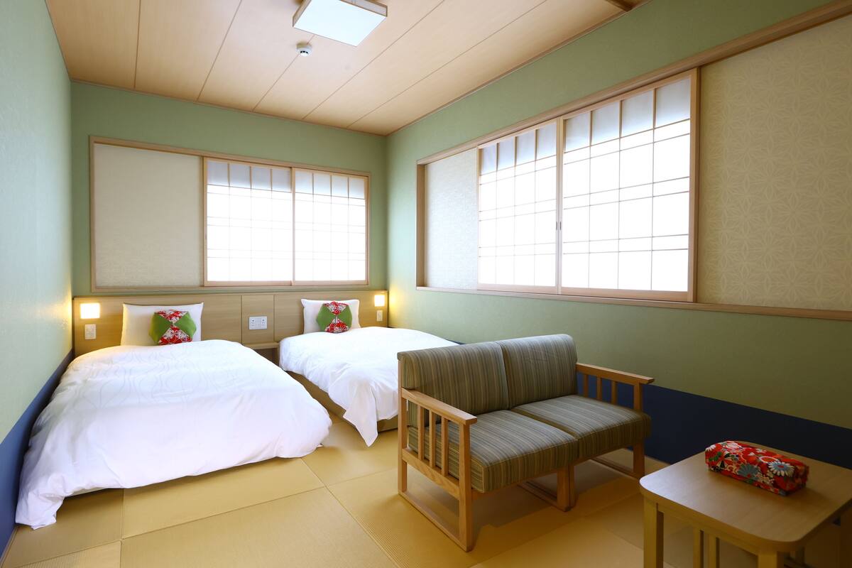 Ein Hotelzimmer mit zwei Betten, einem Sofa, einem kleinen Tisch und großen Fenstern mit traditionellen Schiebetüren.