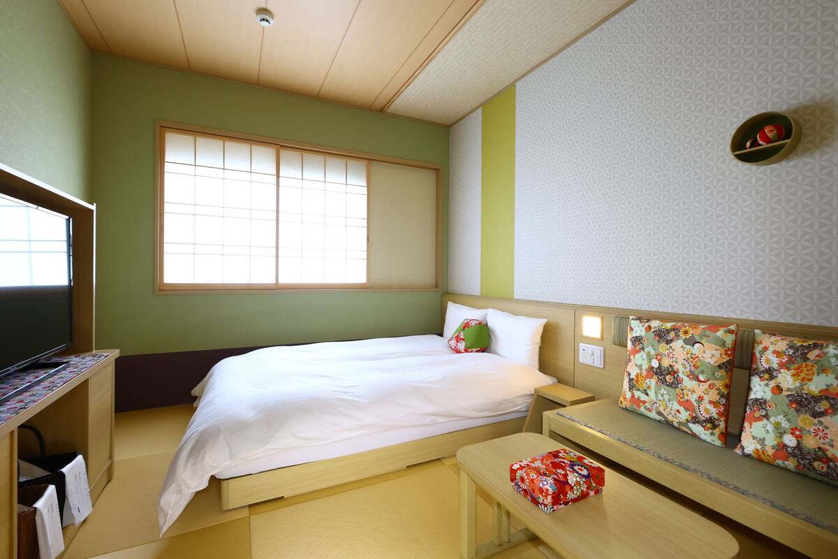 Ein Hotelzimmer mit Bett, Fernseher, einer kleinen Bank und einem Fenster.