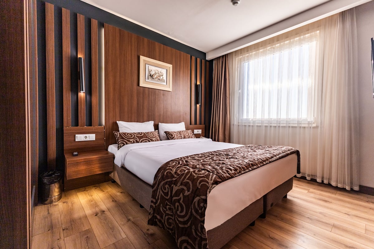 Una habitació d'hotel moderna amb un llit gran, panells de fusta, un quadre amb marc a la paret i una finestra amb cortines de tela fina.
