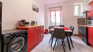 Een compacte keuken met een wasmachine, een koffiezetapparaat en een eettafel met stoelen.