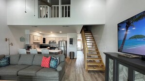 Amplia sala de estar con un sofá gris, una escalera de madera y una cocina al fondo.