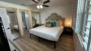 Un dormitorio con cama, ventilador de techo, mesita de noche, ventana con persianas y puerta.