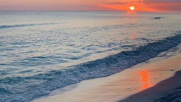 Una playa con un atardecer sobre el océano, olas acariciando suavemente la orilla y un cielo despejado.