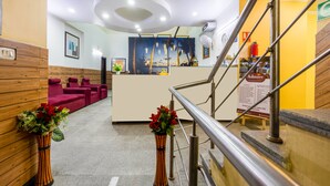 A reception area with a wooden reception desk, a seating area with red couches, and a staircase with metal railings. - Hotel Diamond Stay by ASPL (Noida)
