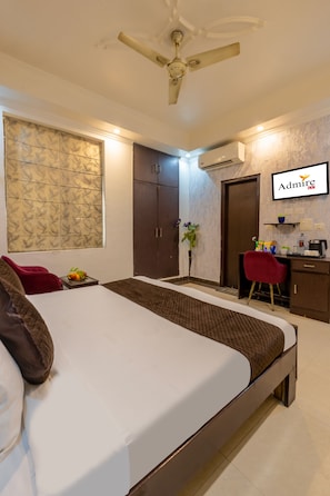 A hotel room with a bed, a desk, a chair, a ceiling fan, an air conditioning unit, and a wall-mounted TV.