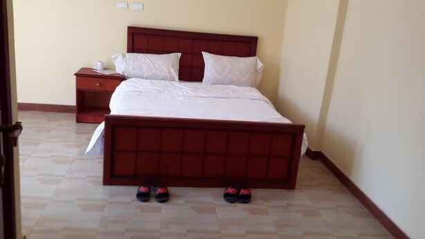 A bedroom with a large bed, a nightstand, and a pair of slippers on the floor. - 20/20 Guest House (Addis Ababa)