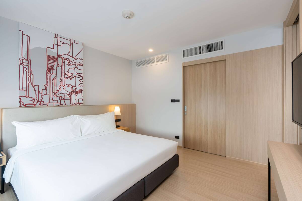 A modern hotel room with a large bed, a nightstand, a wall-mounted artwork, a wooden wardrobe, and a television.
