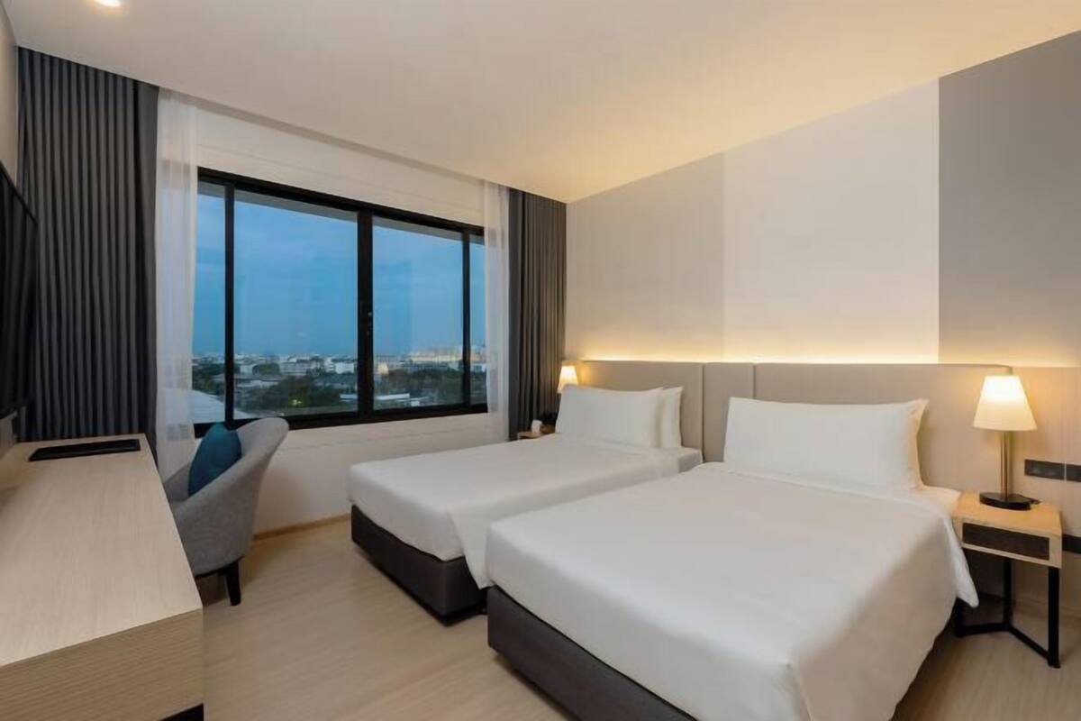 A hotel room with two beds, a desk, a chair, and a large window offering a city view.