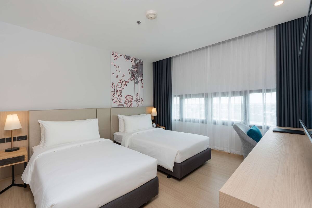 A hotel room with two beds, a desk, a chair, and a large window with curtains.