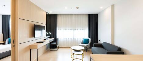 A modern hotel room with a wooden desk, a flat-screen TV, a sofa, and a dining table.